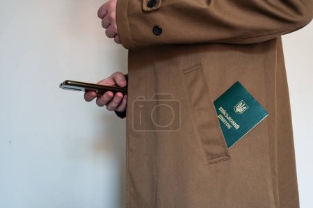Photo for Translation: "military id". Man in coat with smartphone in hand and army document for rookie, veteran, soldier in pocket on white background - Royalty Free Image