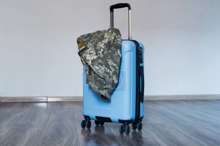Photo for Travel blue suitcase stands on wooden floor with ukrainian military pixel uniform jacket on it - Royalty Free Image