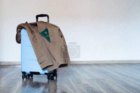 Photo for Translation: "military id". Travel blue suitcase stands on wooden floor with coat. Military green doc sticking out from pocket - Royalty Free Image