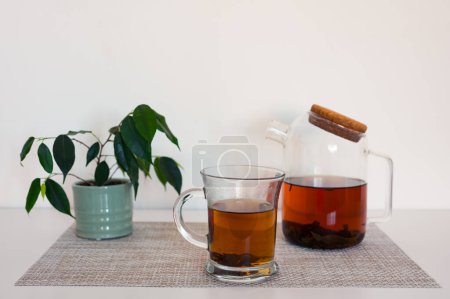 Photo for Black tea in a glass cup and glass teapot on a napkin and green plant on white table with blured white background - Royalty Free Image