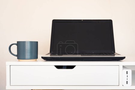 Photo for Empty open laptop with mockup and black screen on white desk table. Cup with coffee. Charger. Minimalist workspace - Royalty Free Image