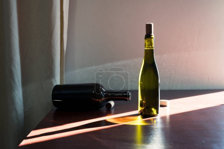 Photo for Green glass bottle of wine with cork and opened dark bottle on table in sun ray. Decor winery cellar bar interior photo - Royalty Free Image