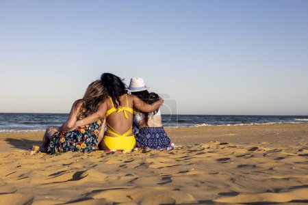 Three girls from behind hugging and sitting on the beach