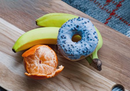 one glazed violet donut, peeled fresh juicy mandarin and two bananas on wooden board. Healthy food concept