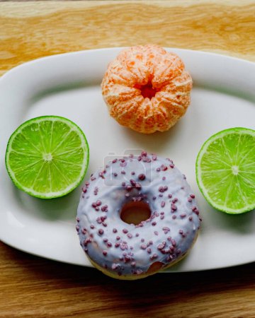 juicy one peeled tangerine, two halfs of green lime and one big round violet glazed donut on white ceramic plate on wooden tray. Fruits concepts. GMO free
