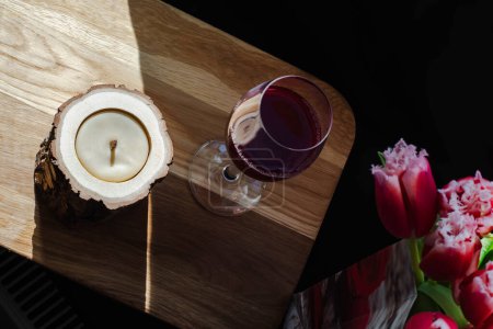 Photo for One glass of aromatic red wine, bouquet of beautiful spring flowers, red tulips in paper present bag and wooden candle on wooden board - Royalty Free Image