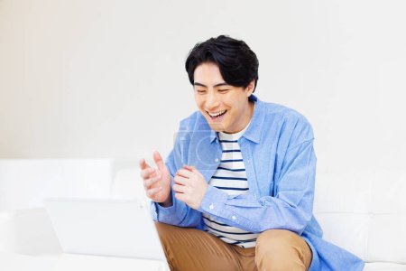 Téléchargez les photos : Portrait de beau jeune Japonais utilisant tablette numérique sur canapé à la maison - en image libre de droit