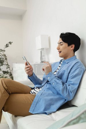 Téléchargez les photos : Jeune asiatique l'homme à l'aide de téléphone mobile tandis que assis sur canapé - en image libre de droit