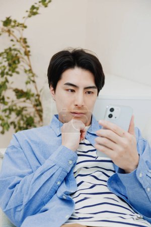 Téléchargez les photos : Portrait de beau jeune Japonais utilisant smartphone sur canapé à la maison - en image libre de droit