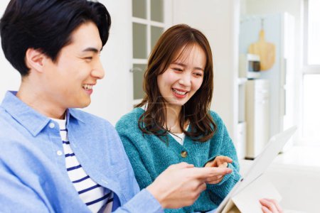 Téléchargez les photos : Jeune couple japonais assis sur le canapé à la maison et en utilisant la tablette numérique ensemble - en image libre de droit