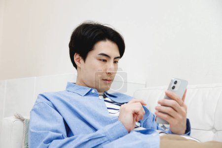 Téléchargez les photos : Portrait de beau jeune Japonais utilisant smartphone sur canapé à la maison - en image libre de droit