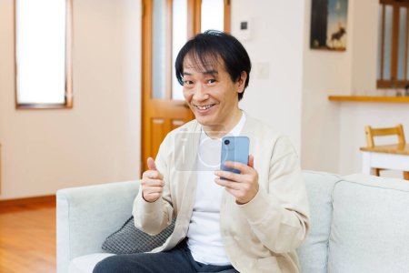 Téléchargez les photos : Asiatique senior homme sourire à la maison à l'aide d 'un téléphone mobile montrant pouce vers le haut - en image libre de droit