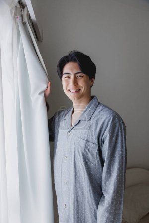 Téléchargez les photos : Portrait de beau jeune Japonais en pyjama debout près de la fenêtre à la maison - en image libre de droit
