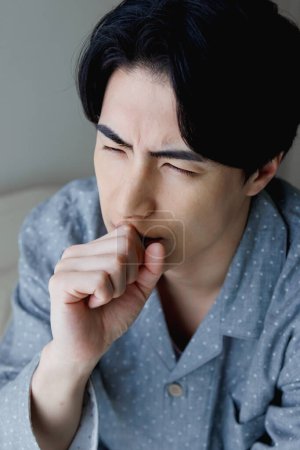 Photo for Portrait of handsome young Japanese man having cough while sitting on bed - Royalty Free Image