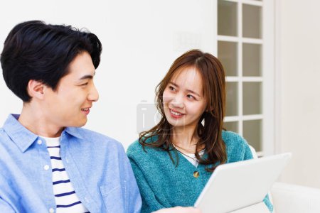 Téléchargez les photos : Jeune couple japonais assis sur le canapé à la maison et en utilisant la tablette numérique ensemble - en image libre de droit