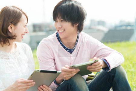 Téléchargez les photos : Couple de jeunes étudiants japonais heureux utilisant tablette numérique passer du temps ensemble sur la nature - en image libre de droit