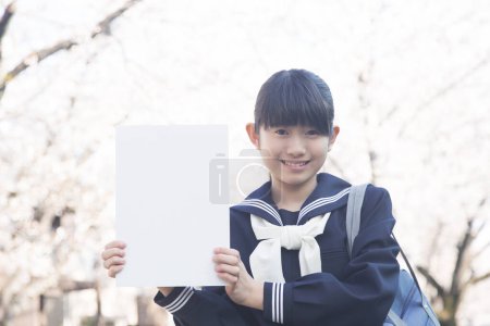 Téléchargez les photos : Étudiant japonais souriant tenant une affiche blanche vierge sur la rue avec des cerisiers en fleurs - en image libre de droit