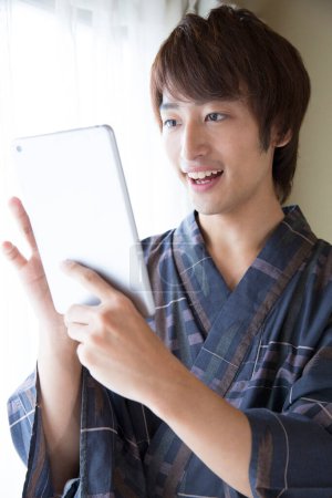 Téléchargez les photos : Portrait de beau jeune homme japonais en yukata traditionnel à l'aide d'une tablette numérique dans un hôtel de source chaude ethnique - en image libre de droit