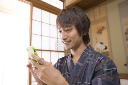 retrato de joven japonés guapo en yukata tradicional usando teléfono inteligente en hotel de aguas termales étnicas