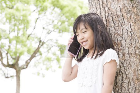 Photo for Cute Japanese girl using smartphone in summer park, daytime portrait of funny kid - Royalty Free Image