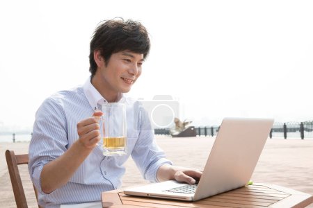 Téléchargez les photos : Jeune homme asiatique boire de la bière et de travailler sur ordinateur portable sur la plage - en image libre de droit