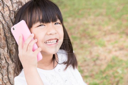 Photo for Cute Japanese girl using smartphone in summer park, daytime portrait of funny kid - Royalty Free Image