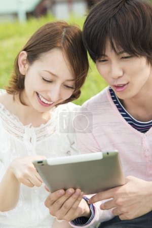 Photo for Couple of happy young japanese students using digital tablet spending time together on nature - Royalty Free Image