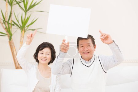 Foto de Retrato de feliz pareja de ancianos mostrando el signo de papel blanco en blanco en la habitación - Imagen libre de derechos