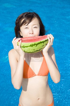 Photo for Young Japanese woman in colorful swimsuit eating watermelon close to pool - Royalty Free Image