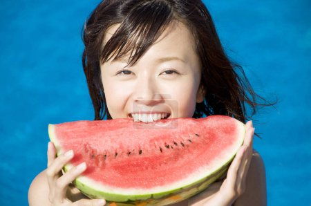 Téléchargez les photos : Jeune femme japonaise en maillot de bain coloré mangeant pastèque près de la piscine - en image libre de droit