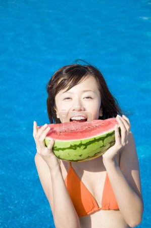 Photo for Young Japanese woman in colorful swimsuit eating watermelon close to pool - Royalty Free Image