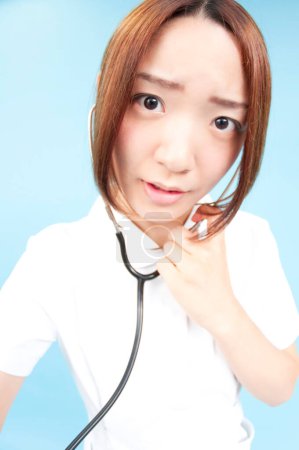 Téléchargez les photos : Portrait grand angle de belle jeune infirmière japonaise avec stéthoscope en uniforme sur fond bleu - en image libre de droit