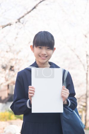 Téléchargez les photos : Étudiant japonais souriant tenant une affiche blanche vierge sur la rue avec des cerisiers en fleurs - en image libre de droit