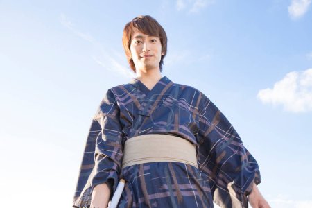 Téléchargez les photos : Portrait de jeune homme japonais beau en yukata traditionnel sur fond de ciel bleu - en image libre de droit