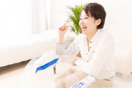 Foto de Mujer asiática con control remoto comiendo un bocadillo en casa - Imagen libre de derechos