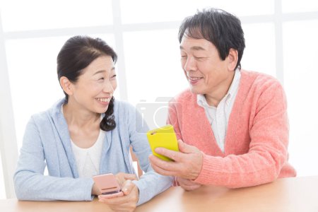 Photo for Senior asian couple reading news on phones at home - Royalty Free Image