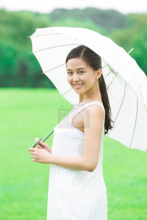 Foto de Mujer japonesa joven con paraguas blanco caminando en el verde parque de verano. Retrato diurno de una joven vestida de blanco - Imagen libre de derechos