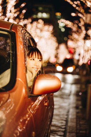 Photo for Young Japanese woman in autumnal clothes sitting in modern car, blurred illuminated city background - Royalty Free Image