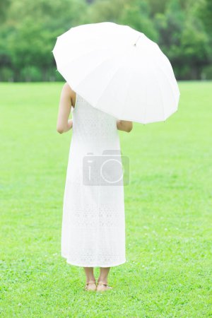 Foto de Mujer japonesa joven con paraguas blanco caminando en el verde parque de verano. Retrato diurno de una joven vestida de blanco - Imagen libre de derechos