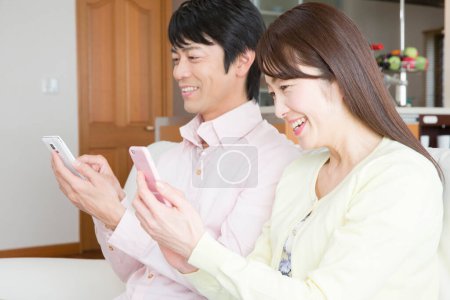 Téléchargez les photos : Heureux asiatique couple en utilisant smartphones sur canapé - en image libre de droit