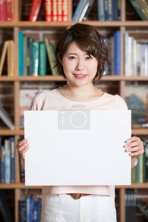 Foto de Sonriente mujer asiática con blanco en blanco signo en la biblioteca - Imagen libre de derechos