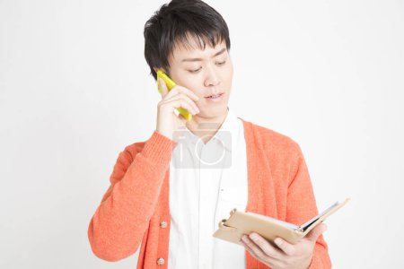 Photo for Asian man rtalking on smartphone and looking in notebook - Royalty Free Image