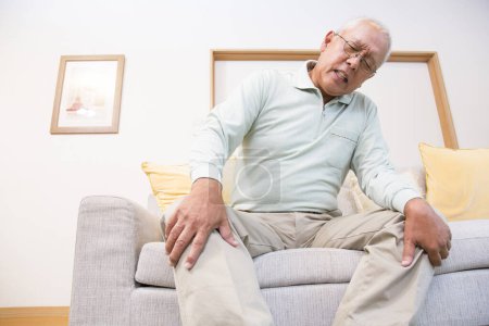 Foto de Senior asiático hombre sentado en sofá y sentir dolor en la rodilla - Imagen libre de derechos