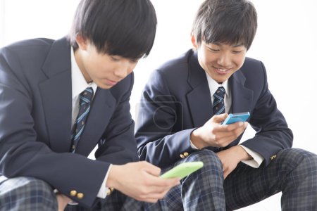 Téléchargez les photos : Deux jeunes garçons asiatiques en uniforme scolaire à l'aide de téléphones mobiles - en image libre de droit