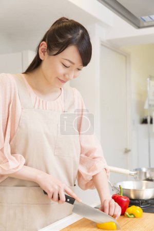 Photo for Beautiful japanese woman in apron cutting bell pepper in kitchen - Royalty Free Image