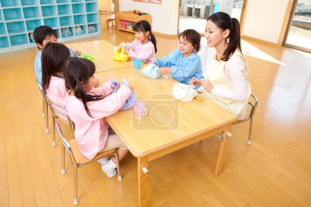 Foto de Maestra de jardín de infantes y niños sentados a la mesa durante la hora del almuerzo - Imagen libre de derechos