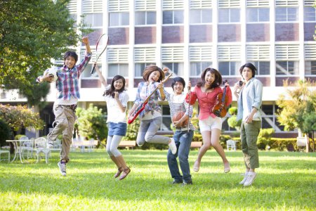 Foto de Feliz grupo de estudiantes de las secciones de música y deporte saltando en el césped verde - Imagen libre de derechos