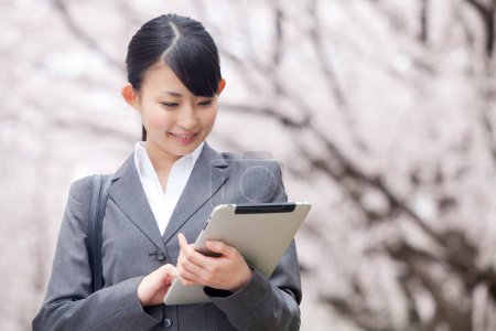Téléchargez les photos : Portrait de belle jeune femme d'affaires japonaise utilisant une tablette numérique dans le parc pendant la saison de floraison sakura - en image libre de droit