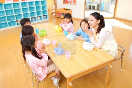 Téléchargez les photos : Professeur de maternelle et les enfants assis à table pendant l'heure du déjeuner - en image libre de droit