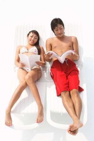 Téléchargez les photos : Jeune couple asiatique avec des livres sur les chaises longues sur une plage - en image libre de droit